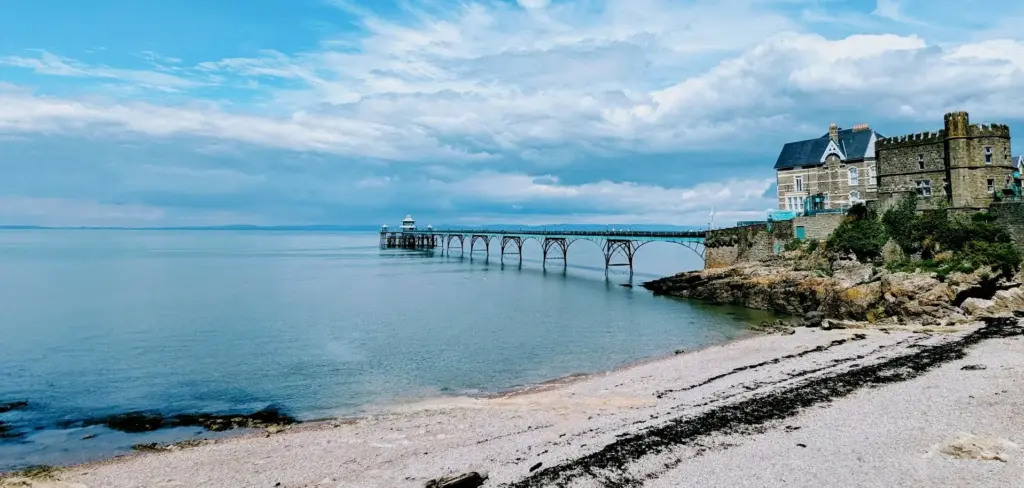Scenic view of Clevedon Pier stretching over calm sea waters, with a historic stone building on a rocky coast under a partly cloudy blue sky.
