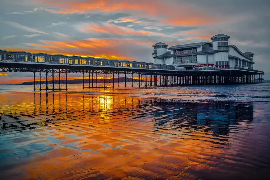 The Grand Pier at Weston-super-Mare captured at sunset, reflecting vivid orange and purple hues on the wet sandy beach.