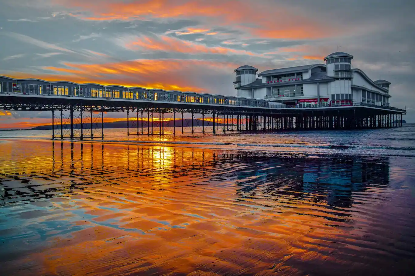 The Grand Pier at Weston-super-Mare captured at sunset, reflecting vivid orange and purple hues on the wet sandy beach.